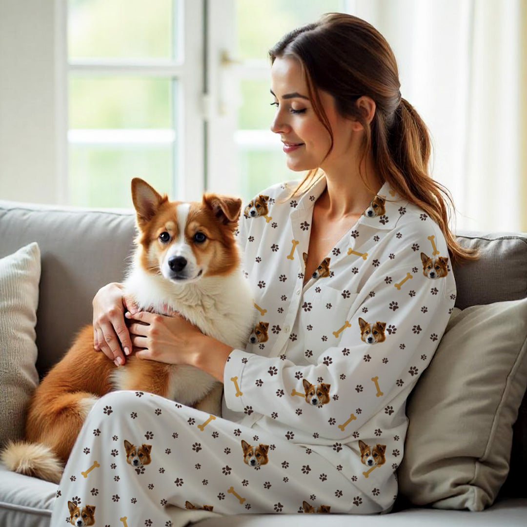 Femme portant un pyjama blanc personnalisé avec ne tête de chien, des empreintes de pattes et un motif os, tenant un chien brun et blanc sur le canapé